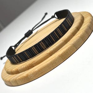 Peut inclure: Un bracelet noir à perles avec des perles rectangulaires marron foncé et clair alternées. Le bracelet est posé sur une surface en bois brun clair. Le bracelet a une fermeture à cordon réglable noire.