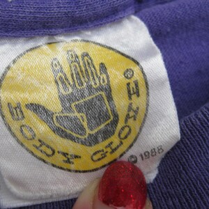 May include: Close-up of a vintage purple t-shirt tag. The tag is white with a yellow oval featuring a black hand graphic and the words "BODY GLOVE". The year "1988" is printed below the graphic.