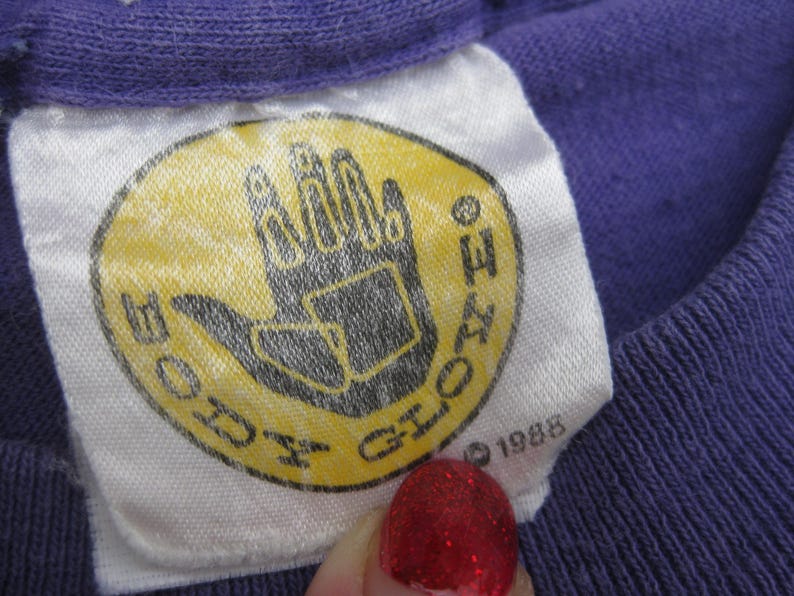 May include: Close-up of a vintage purple t-shirt tag. The tag is white with a yellow oval featuring a black hand graphic and the words "BODY GLOVE". The year "1988" is printed below the graphic.