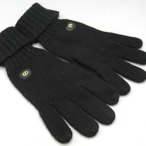 May include: A pair of black knit gloves featuring a ribbed cuff design and a small circular black leather patch on the back of each hand. Each patch contains a gold-toned metallic emblem with the letter F centered within the circular detail.