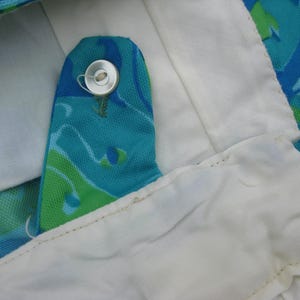 May include: Close-up of a vintage garment detail. The image shows a buttoned closure with a silver button on a turquoise fabric with a green and blue pattern. The garment has a white lining.