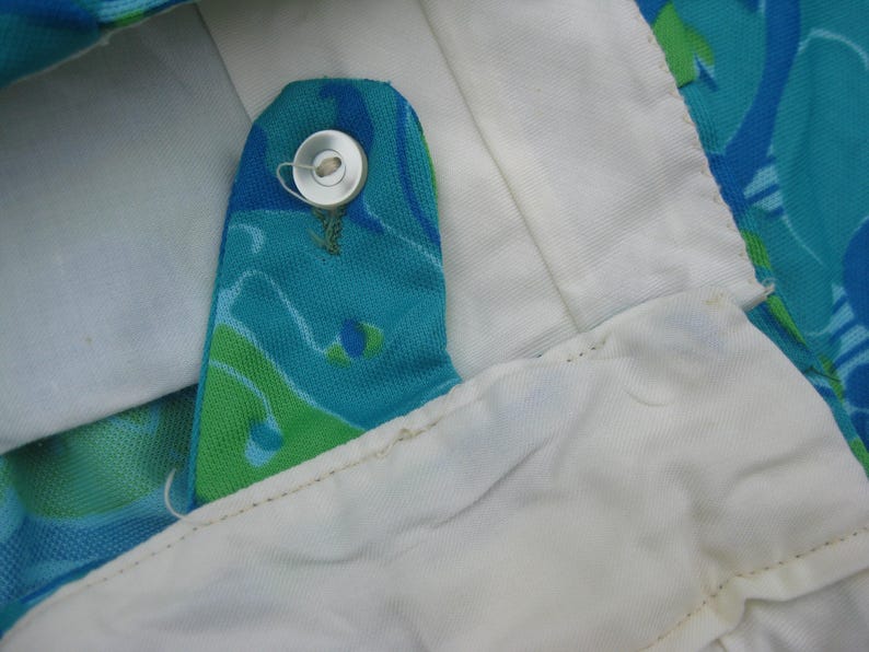 May include: Close-up of a vintage garment detail. The image shows a buttoned closure with a silver button on a turquoise fabric with a green and blue pattern. The garment has a white lining.