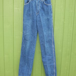 May include: Blue denim jeans with a high waist and straight leg. The jeans have classic five-pocket styling, a button and zipper closure, and belt loops. The jeans are hanging on a white hanger against a green background.