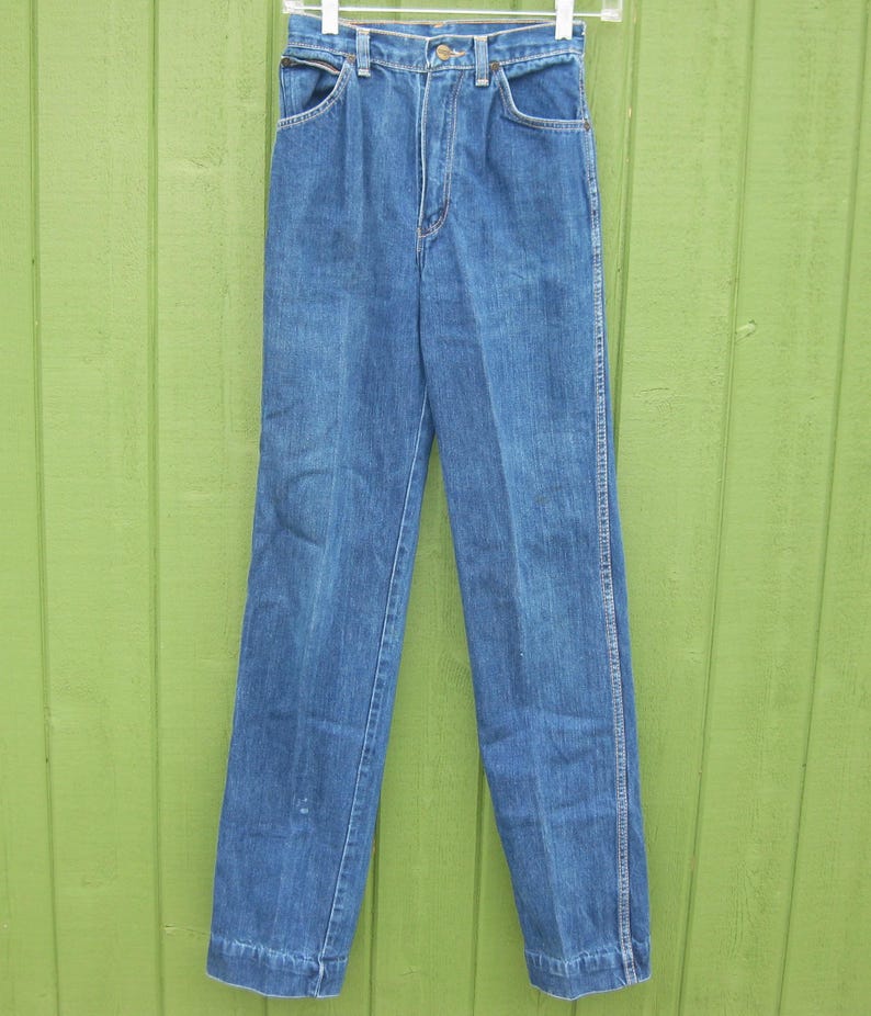 May include: Blue denim jeans with a high waist and straight leg. The jeans have classic five-pocket styling, a button and zipper closure, and belt loops. The jeans are hanging on a white hanger against a green background.