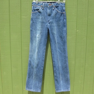 May include: Blue denim jeans with a classic five-pocket design. The jeans are straight-legged and have a faded wash, with visible wear marks. They are hanging on a metal hanger against a green background.