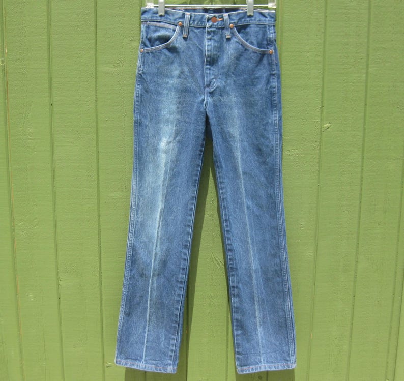 May include: Blue denim jeans with a classic five-pocket design. The jeans are straight-legged and have a faded wash, with visible wear marks. They are hanging on a metal hanger against a green background.