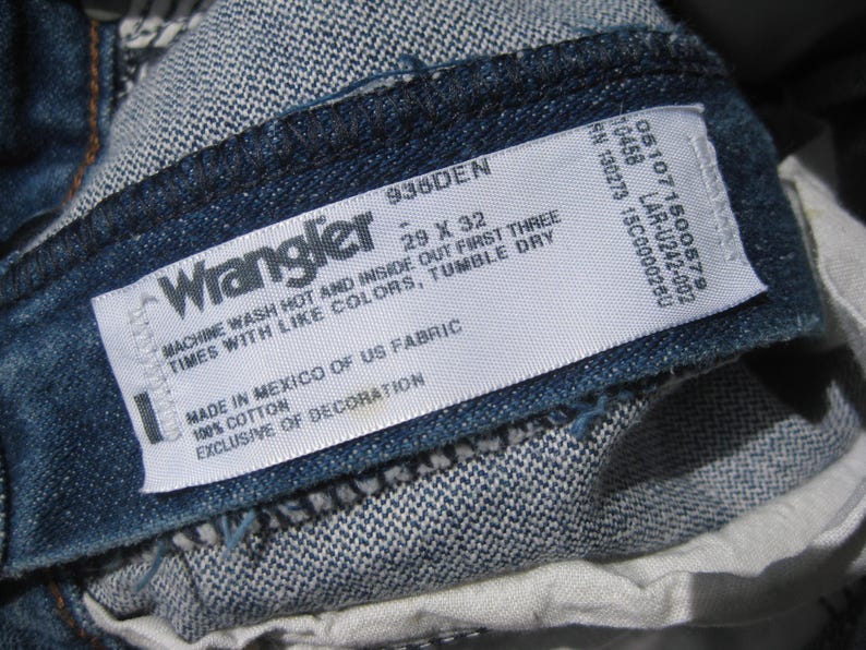 May include: Close-up of a denim Wrangler jeans label. The label displays the brand name, size 29 x 32, and care instructions. The jeans are made of 100% cotton and manufactured in Mexico.
