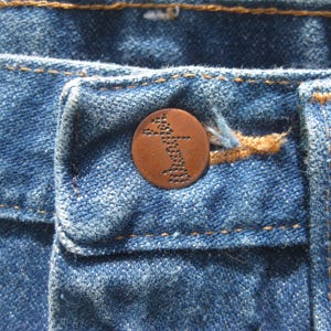 May include: Close-up of blue denim jeans with a copper button. The button features a detailed design. The jeans have visible stitching in a contrasting orange-brown thread. The fabric shows signs of wear.