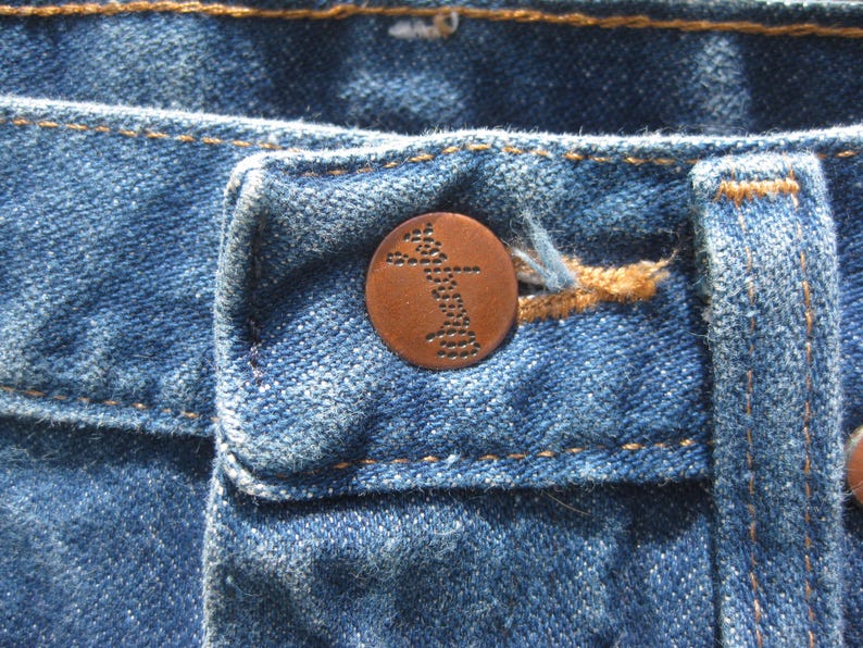 May include: Close-up of blue denim jeans with a copper button. The button features a detailed design. The jeans have visible stitching in a contrasting orange-brown thread. The fabric shows signs of wear.