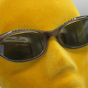Puede incluir: Gafas de sol marrones con forma de ojo de gato y lentes oscuras. Las monturas tienen un aspecto sutil y texturizado, sobre un fondo amarillo. Un estilo cl&aacute;sico de gafas de sol, adecuado para diversas ocasiones.