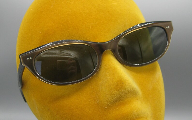 Puede incluir: Gafas de sol marrones con forma de ojo de gato y lentes oscuras. Las monturas tienen un aspecto sutil y texturizado, sobre un fondo amarillo. Un estilo cl&aacute;sico de gafas de sol, adecuado para diversas ocasiones.