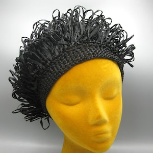 May include: A black woven hat with a textured, fringed brim. The hat is displayed on a yellow mannequin head. The hat's design features a woven band and a fringe of black, ribbon-like material.