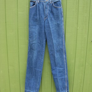 May include: Blue denim jeans with a classic straight leg design. The jeans have a button and zipper closure, front and back pockets, and belt loops. The jeans are hanging on a white hanger against a green background.