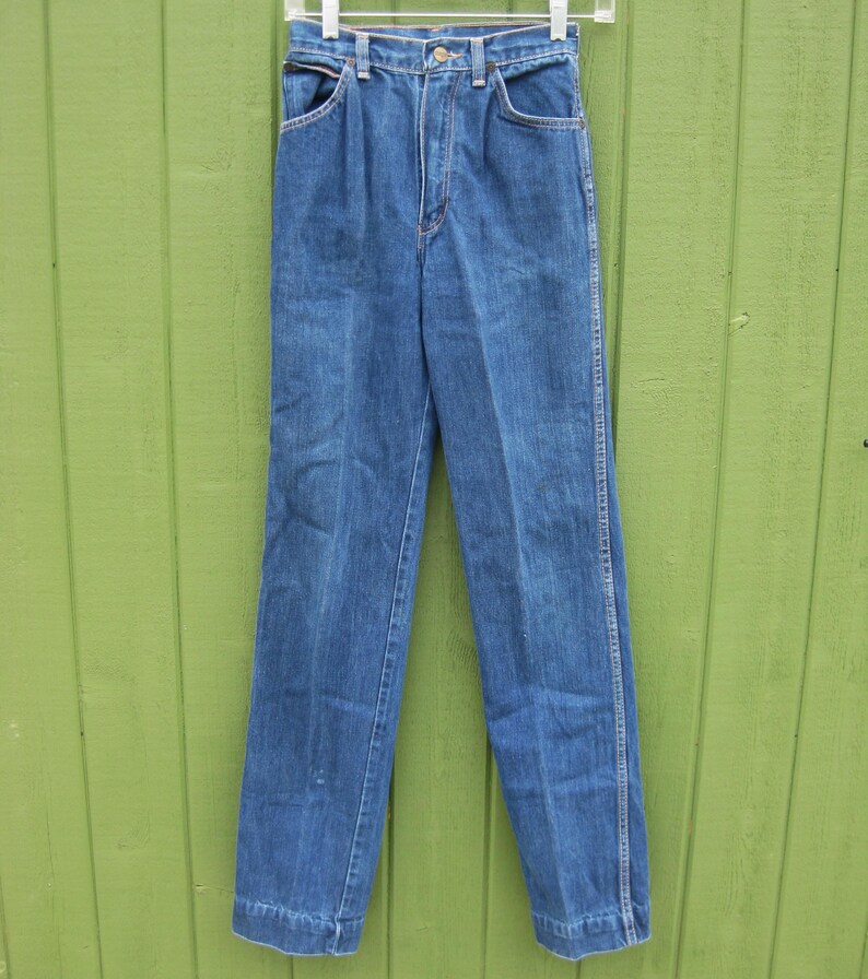 May include: Blue denim jeans with a classic straight leg design. The jeans have a button and zipper closure, front and back pockets, and belt loops. The jeans are hanging on a white hanger against a green background.