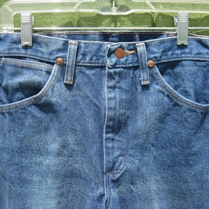 May include: Close-up of blue denim jeans hanging on a clear plastic hanger. The jeans feature classic five-pocket styling with copper rivets and a button closure. The denim has a faded, worn appearance.