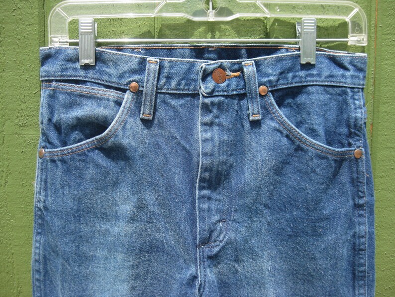 May include: Close-up of blue denim jeans hanging on a clear plastic hanger. The jeans feature classic five-pocket styling with copper rivets and a button closure. The denim has a faded, worn appearance.