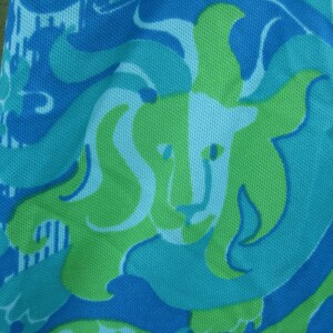 May include: Close-up of a fabric with a repeating lion design. The pattern features a lion's head in light green, surrounded by swirling shapes in shades of blue and turquoise. The fabric has a textured appearance.