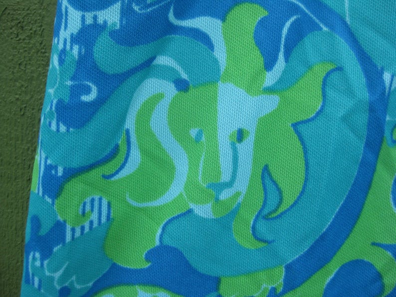 May include: Close-up of a fabric with a repeating lion design. The pattern features a lion's head in light green, surrounded by swirling shapes in shades of blue and turquoise. The fabric has a textured appearance.