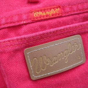 Peut inclure: Gros plan de la poche arri&egrave;re d'un jean Wrangler rose vif. Le logo Wrangler est visible sur une petite &eacute;tiquette rouge et un patch en cuir marron. Les coutures sont de couleur orange contrastante.
