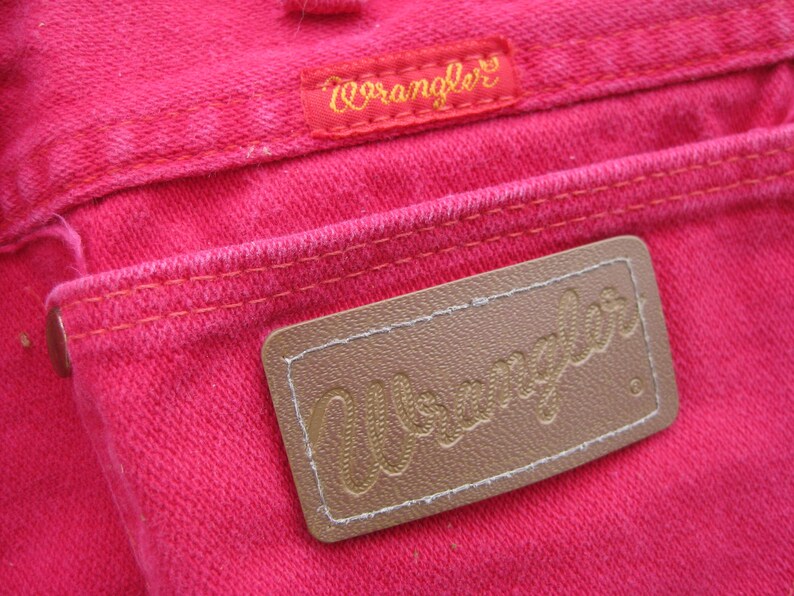 Peut inclure: Gros plan de la poche arri&egrave;re d'un jean Wrangler rose vif. Le logo Wrangler est visible sur une petite &eacute;tiquette rouge et un patch en cuir marron. Les coutures sont de couleur orange contrastante.