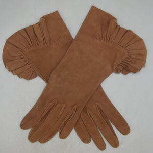 May include: A pair of tan suede gloves with a ruffled cuff design. The gloves are long, extending past the wrist, and feature detailed stitching along the fingers and seams. The gloves are laid out on a light background.