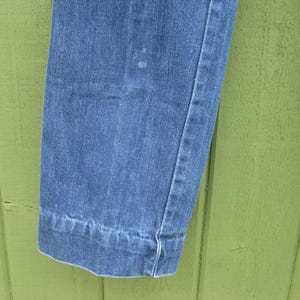May include: Close-up of a section of blue denim jeans. The image shows the hem of the jeans, with a visible seam and a slight cuff. The denim has a classic, faded appearance, with subtle variations in color.
