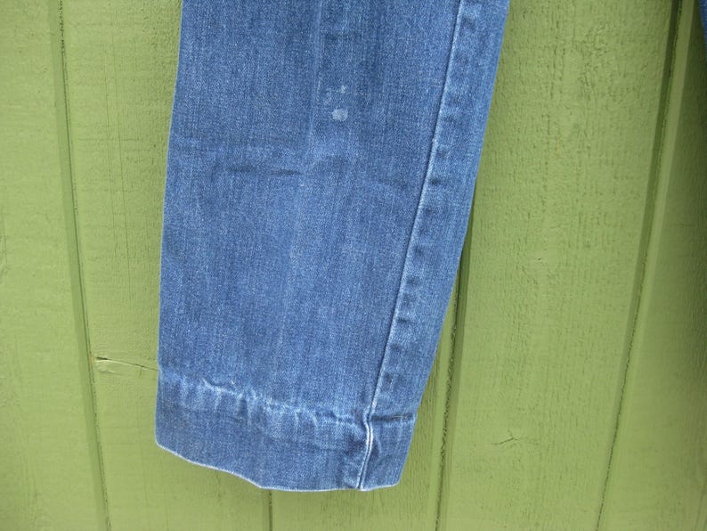 May include: Close-up of a section of blue denim jeans. The image shows the hem of the jeans, with a visible seam and a slight cuff. The denim has a classic, faded appearance, with subtle variations in color.