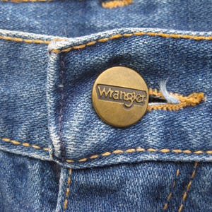 May include: Close-up of blue denim jeans with a brass button. The button is embossed with the brand name "Wrangler". The jeans have orange stitching along the seams and around the buttonhole.