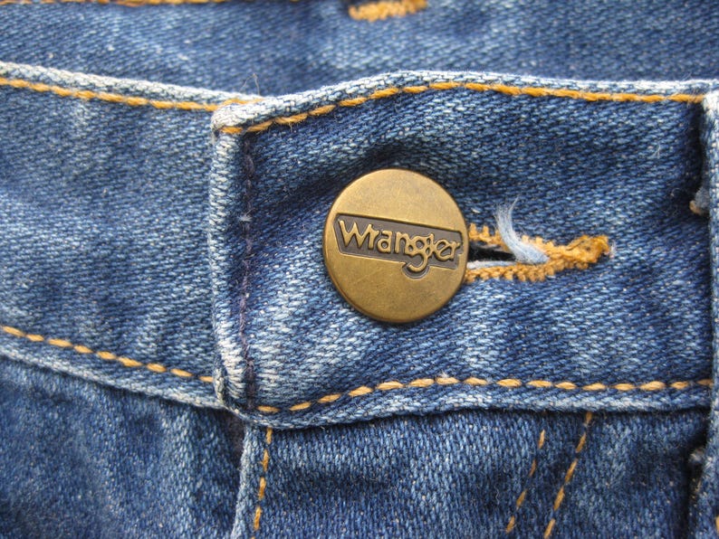 May include: Close-up of blue denim jeans with a brass button. The button is embossed with the brand name "Wrangler". The jeans have orange stitching along the seams and around the buttonhole.
