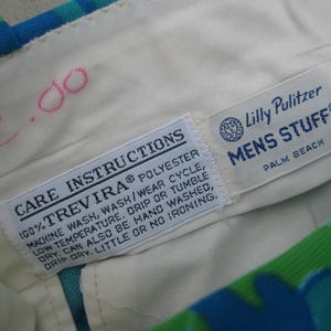 May include: Close-up of the interior waistband of a pair of patterned pants. The cream-colored fabric features two sewn-in tags with care instructions and the "Lilly Pulitzer MENS STUFF" logo. The pants have a blue, green, and white floral pattern.