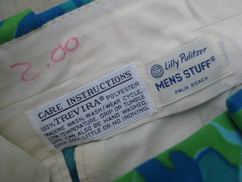 May include: Close-up of the interior waistband of a pair of patterned pants. The cream-colored fabric features two sewn-in tags with care instructions and the "Lilly Pulitzer MENS STUFF" logo. The pants have a blue, green, and white floral pattern.