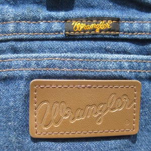 May include: Close-up of blue denim fabric with brown stitching. Features two Wrangler brand labels: a small black label with gold text and a larger brown leather label with embossed text.