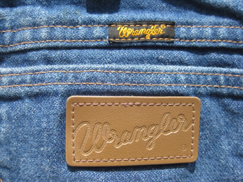 May include: Close-up of blue denim fabric with brown stitching. Features two Wrangler brand labels: a small black label with gold text and a larger brown leather label with embossed text.