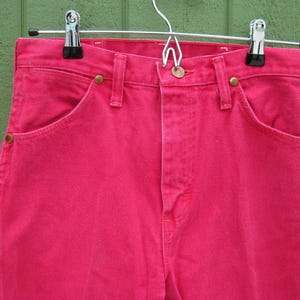 Peut inclure: Un jean en denim rose vif avec une coupe classique &agrave; cinq poches, une fermeture boutonn&eacute;e et des rivets en laiton. Le jean est suspendu &agrave; un cintre m&eacute;tallique sur un fond vert. Gros plan sur les d&eacute;tails du v&ecirc;tement.