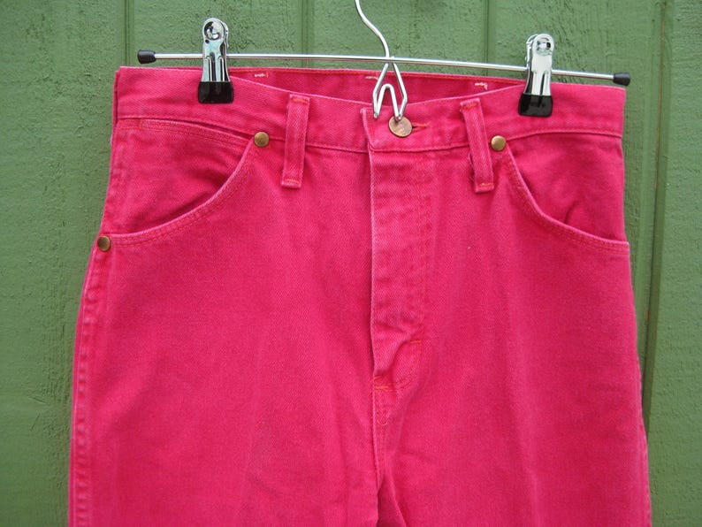 Peut inclure: Un jean en denim rose vif avec une coupe classique &agrave; cinq poches, une fermeture boutonn&eacute;e et des rivets en laiton. Le jean est suspendu &agrave; un cintre m&eacute;tallique sur un fond vert. Gros plan sur les d&eacute;tails du v&ecirc;tement.