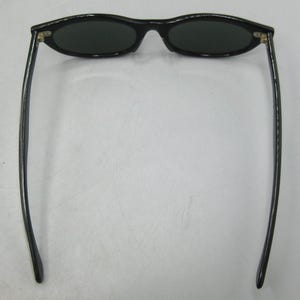 Puede incluir: Gafas de sol ovaladas negras con lentes oscuras. Las gafas tienen una montura elegante y curvada y patillas largas y delgadas. Las lentes son de color verde oscuro. Se muestran desde una perspectiva superior.