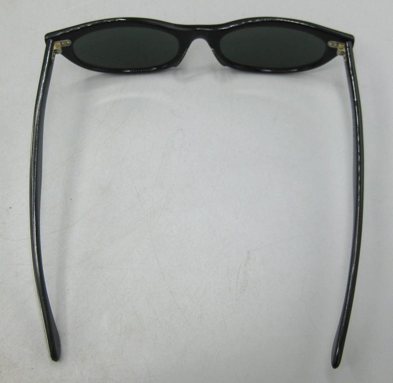 Puede incluir: Gafas de sol ovaladas negras con lentes oscuras. Las gafas tienen una montura elegante y curvada y patillas largas y delgadas. Las lentes son de color verde oscuro. Se muestran desde una perspectiva superior.