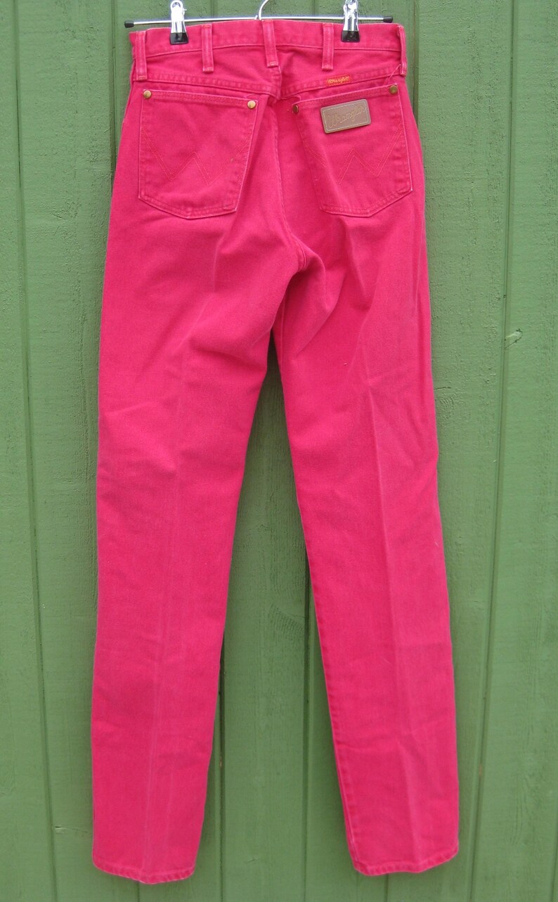Peut inclure: Un jean en denim rose vif suspendu &agrave; un cintre en m&eacute;tal. Le jean a deux poches arri&egrave;re, un patch en cuir marron et des rivets dor&eacute;s. Le jean est d'une couleur vive et unie et semble &ecirc;tre vintage.