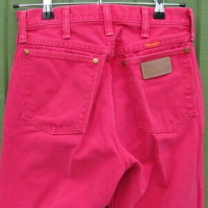 Peut inclure: Gros plan sur l'arri&egrave;re d'un jean Wrangler rose vif. Le jean a deux poches arri&egrave;re avec le logo Wrangler et une petite &eacute;tiquette au-dessus de la poche droite. Le jean est suspendu &agrave; un cintre en m&eacute;tal.