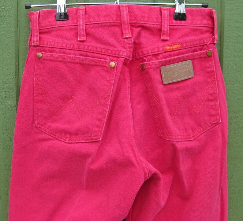 Peut inclure: Gros plan sur l'arri&egrave;re d'un jean Wrangler rose vif. Le jean a deux poches arri&egrave;re avec le logo Wrangler et une petite &eacute;tiquette au-dessus de la poche droite. Le jean est suspendu &agrave; un cintre en m&eacute;tal.
