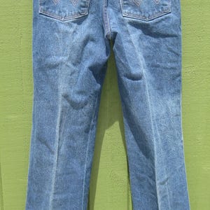May include: Blue denim jeans with a classic five-pocket design. Features include copper-coloured stitching, a leather patch with the brand name, and the iconic 'W' stitching on the back pockets. The jeans are hanging on a metal hanger.