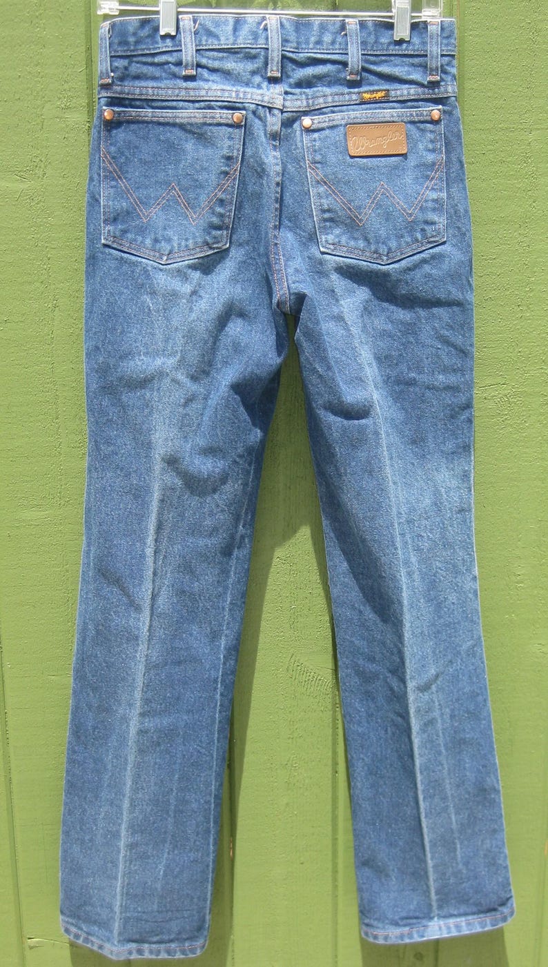 May include: Blue denim jeans with a classic five-pocket design. Features include copper-coloured stitching, a leather patch with the brand name, and the iconic 'W' stitching on the back pockets. The jeans are hanging on a metal hanger.