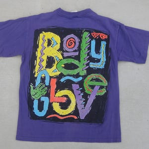 May include: A purple t-shirt featuring a colorful graphic design that reads "Body Glove" in a variety of bright colors. The design is set against a black background. The shirt has short sleeves and a crew neck.