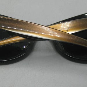 Puede incluir: Gafas de sol negras de ojo de gato con patillas doradas. Las gafas tienen un dise&ntilde;o cl&aacute;sico con un aspecto elegante y moderno. Las patillas est&aacute;n cruzadas, mostrando los detalles de las bisagras y la forma de la montura. Las lentes son oscuras.