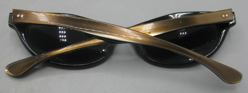 Puede incluir: Gafas de sol negras de ojo de gato con patillas doradas. Las gafas tienen un dise&ntilde;o cl&aacute;sico con un aspecto elegante y moderno. Las patillas est&aacute;n cruzadas, mostrando los detalles de las bisagras y la forma de la montura. Las lentes son oscuras.