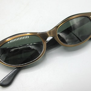 Puede incluir: Gafas de sol vintage con montura dorada y lentes oscuras. Las gafas tienen una forma de ojo de gato cl&aacute;sica con patillas negras. La montura tiene peque&ntilde;os detalles decorativos cerca de las bisagras.