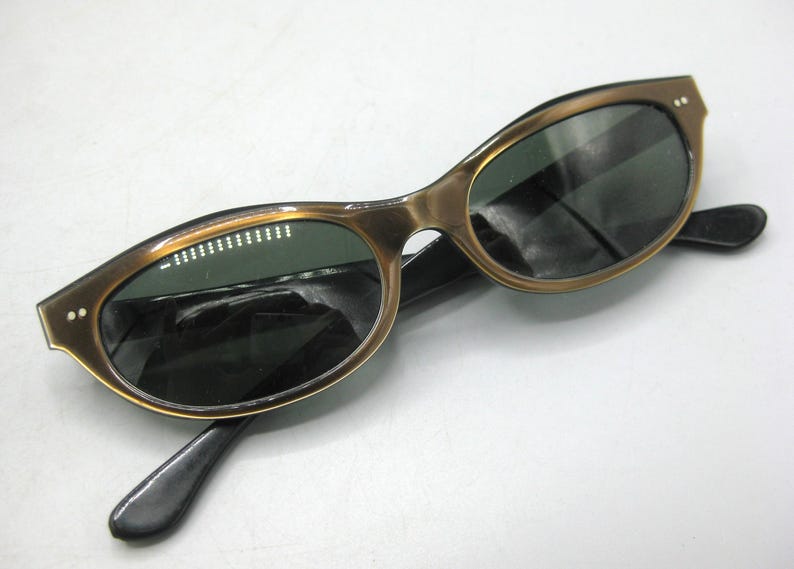 Puede incluir: Gafas de sol vintage con montura dorada y lentes oscuras. Las gafas tienen una forma de ojo de gato cl&aacute;sica con patillas negras. La montura tiene peque&ntilde;os detalles decorativos cerca de las bisagras.