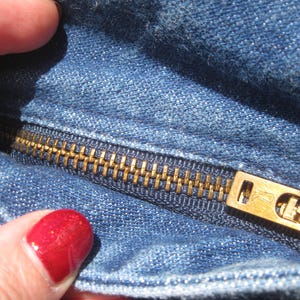 May include: Close-up of blue denim fabric with a brass zipper. The zipper is partially open, showing the teeth and slider. The denim has a classic, worn appearance.