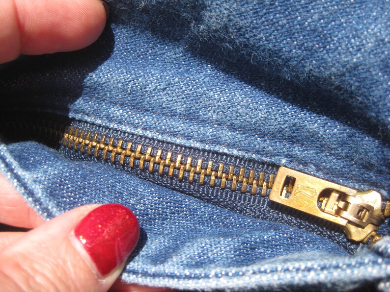 May include: Close-up of blue denim fabric with a brass zipper. The zipper is partially open, showing the teeth and slider. The denim has a classic, worn appearance.