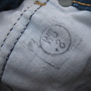 May include: Close-up of the inside of denim jeans, showing the white fabric lining and stitching. A faded circular stamp with the numbers "28" is visible. The jeans are blue.